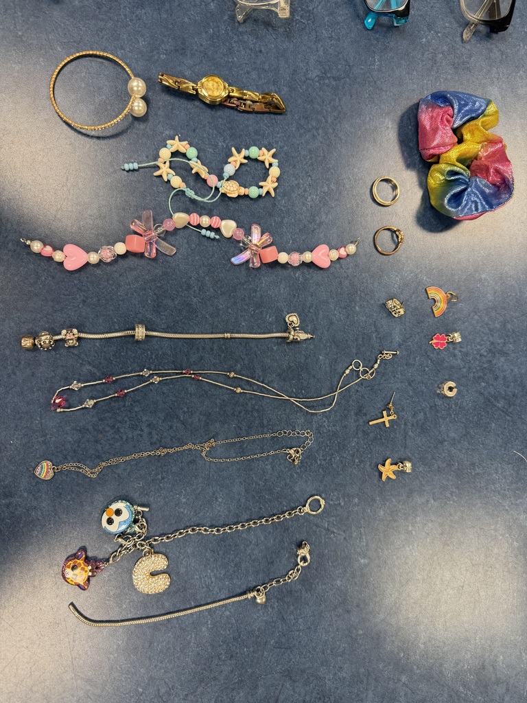 Assorted jewelry on a blue surface, including several beaded bracelets (pink, blue, and starfish themes), silver charm bracelets, delicate necklaces, rings, earrings, a gold watch, and a rainbow scrunchie.