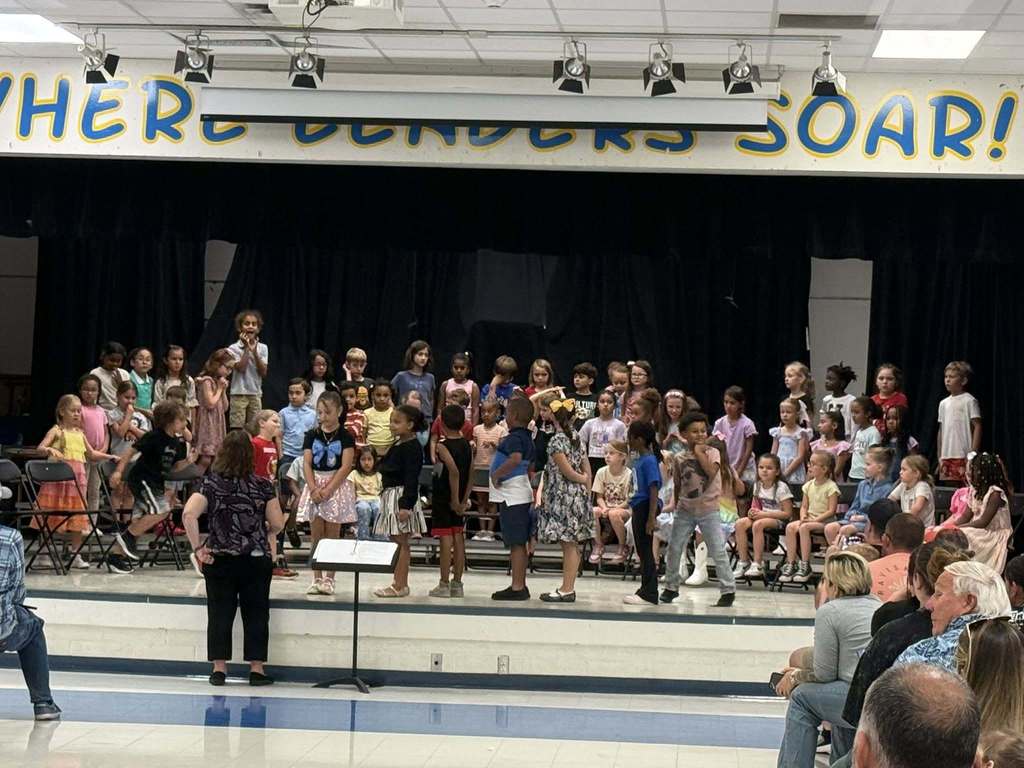 A group of approximately 40 elementary-aged children performs on a stage during a school assembly. They are dressed in colorful casual attire, standing and sitting on risers. A teacher stands with her back to the camera, directing them. Above, a banner reads "WHERE READERS SOAR!" under stage lights.