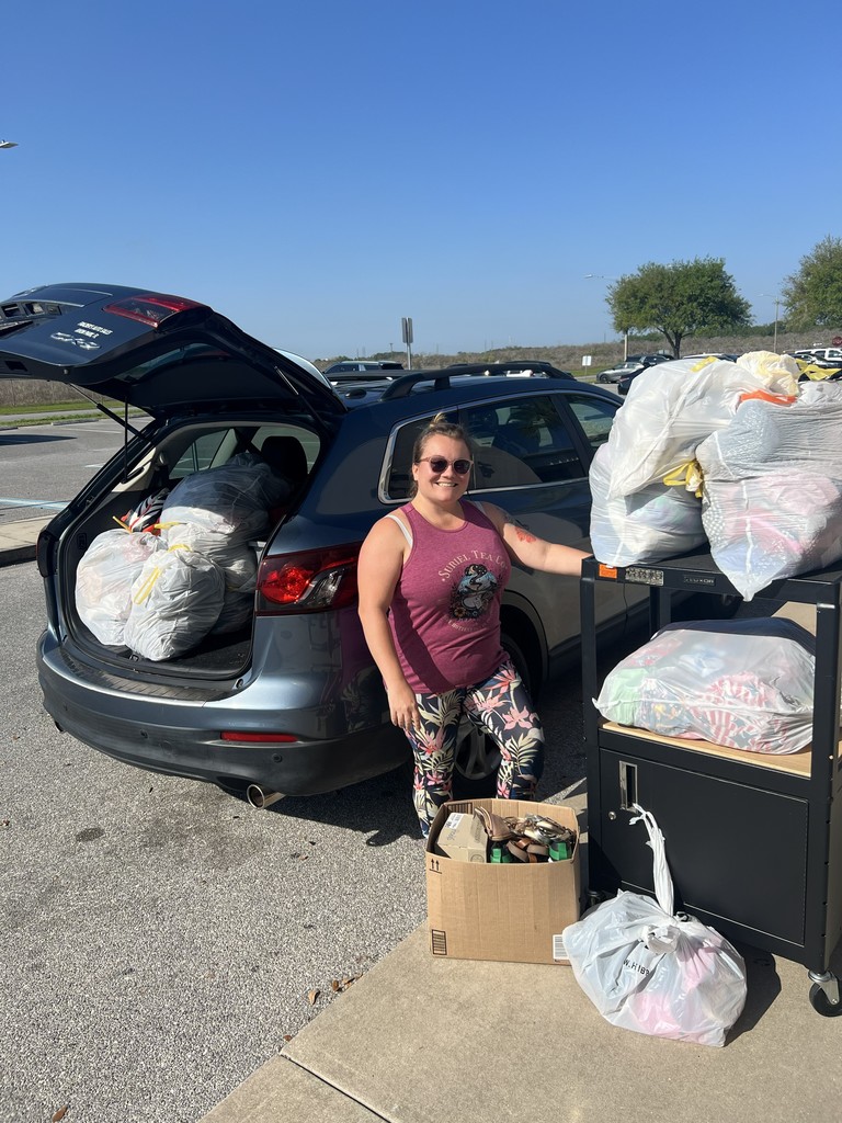 Christa Jones dropping of bagged donations of clothes and shoes for MES students. Bags pictured in back of car and on a cart. Picture taken outside with blue sky and green trees and gray road and sidewalk,