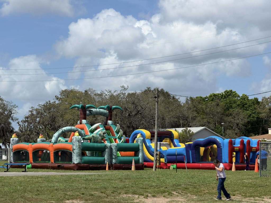  Gemini said An outdoor scene featuring two large, colorful inflatable obstacle courses on a grassy field under a bright, cloudy sky. On the left is a green and orange dinosaur-themed course; on the right is a blue, yellow, and red course. A young girl walks in the foreground.