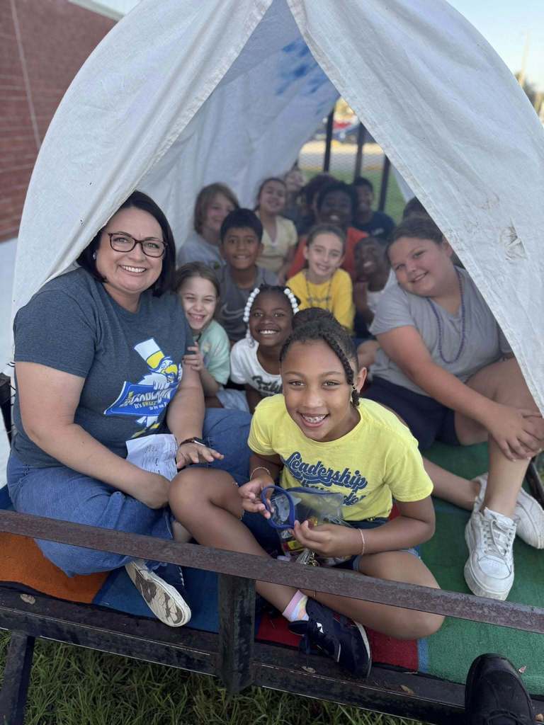 A female teacher with glasses and a gray t-shirt smiles while sitting with a group of approximately ten students inside a makeshift covered wagon. The students are diverse, cheerful, and seated on colorful mats. One student in a yellow shirt sits prominently in the foreground.