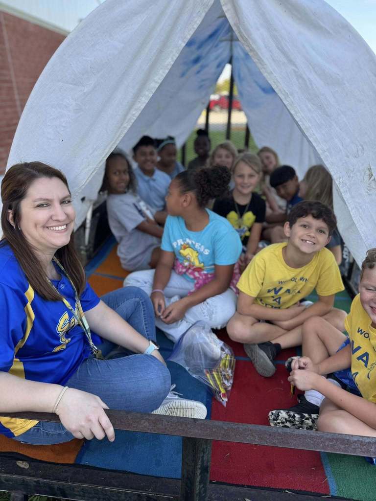 A smiling teacher in a blue athletic shirt sits in the foreground on a trailer. Inside the white fabric canopy behind her, several elementary-aged children sit on colorful rugs, looking toward the camera. One child in a bright blue "Princess" shirt sits near the center.