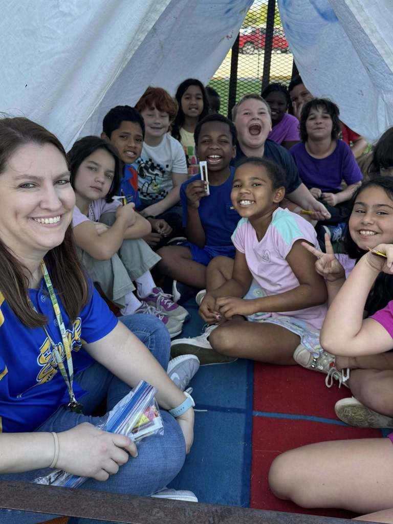 A teacher in a blue jersey smiles, holding a bag of snacks, while sitting on a flatbed trailer. A group of excited children are gathered behind her under a white arched canopy. The children are laughing and making peace signs, seated on blue and red mats.