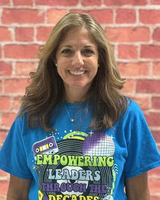 A smiling woman with shoulder-length wavy brown hair stands before a red brick wall. She wears a bright blue t-shirt featuring a retro "Empowering Leaders Through the Decades" graphic with a cassette tape and disco ball. She has a fair complexion, small pearl earrings, and a delicate silver necklace.