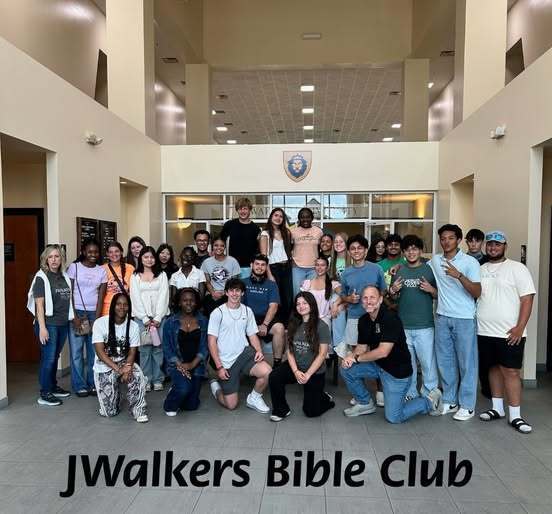 A large, diverse group of students and two adults pose for a group photo in a bright, modern building lobby. They are standing and kneeling in multiple rows under a crest logo. Text at the bottom of the image reads, "JWalkers Bible Club."