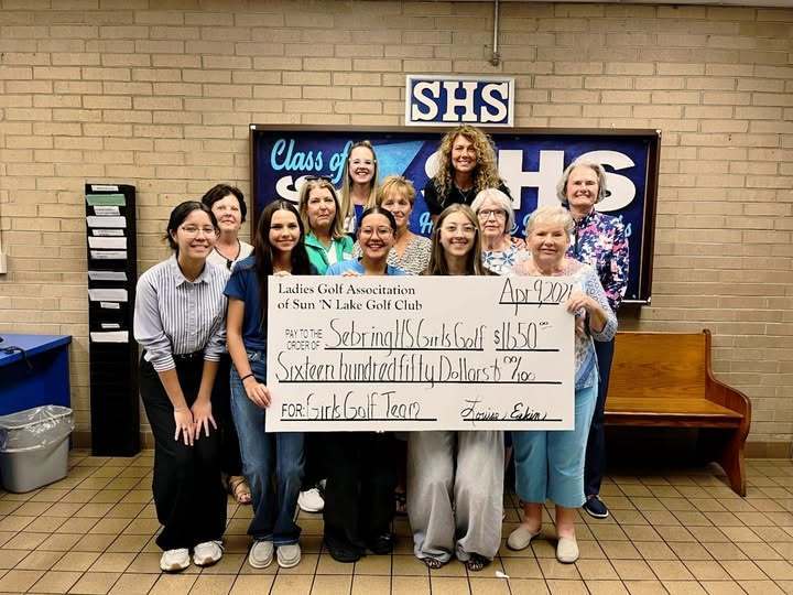 A group of eleven women and students pose indoors, holding a large ceremonial check for $1,650. The check is from the Ladies Golf Association of Sun 'N Lake Golf Club to the Sebring HS Girls Golf team. They stand in front of a brick wall and "SHS" sign.