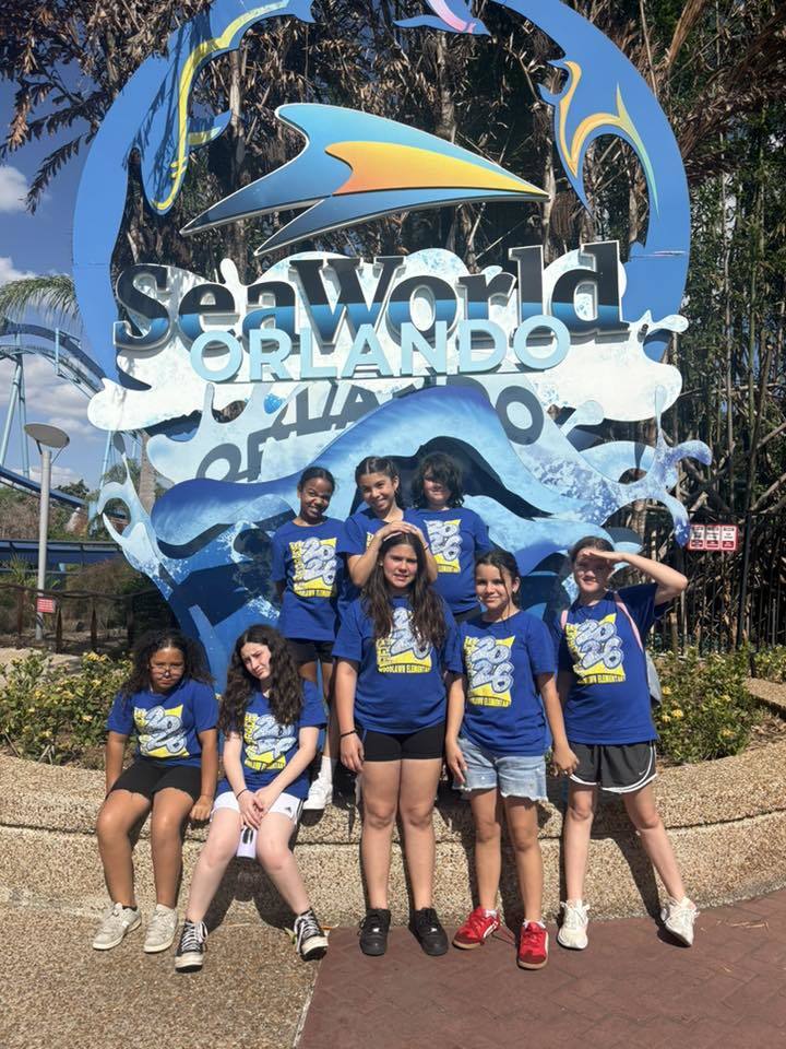  Seven children in matching blue "Class of 2026" t-shirts pose in front of the SeaWorld Orlando entrance sign. The group is arranged in two rows against a large blue and white wave-themed backdrop. A roller coaster track and tropical trees are visible in the sunny background.