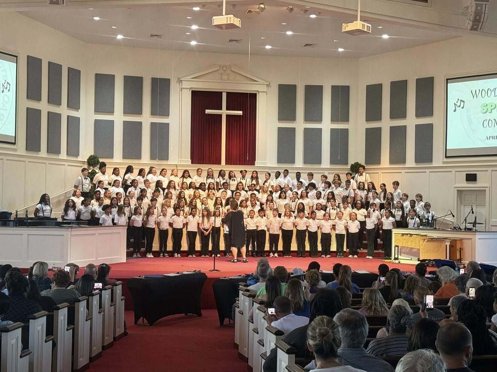 A large youth choir in white shirts and dark pants performs on a tiered stage in a church sanctuary. A conductor stands before them with their back to a seated audience. A white cross hangs against a red curtain in the background, flanked by grey acoustic panels.