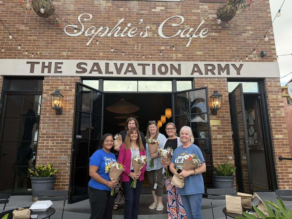 Six women stand smiling in the doorway of "Sophie’s Cafe," part of a brick Salvation Army building. Five women hold small floral bouquets. The storefront features black-framed glass doors, industrial wall lanterns, and string lights. Outdoor cafe tables with brown paper bags are in the foreground.