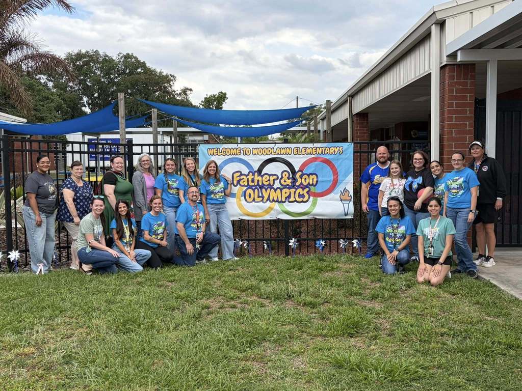 A group of adults, mostly wearing matching blue school spirit shirts, pose outdoors in front of a black fence. They are gathered around a colorful banner that reads "Welcome to Woodlawn Elementary’s Father & Son Olympics," featuring the Olympic rings.