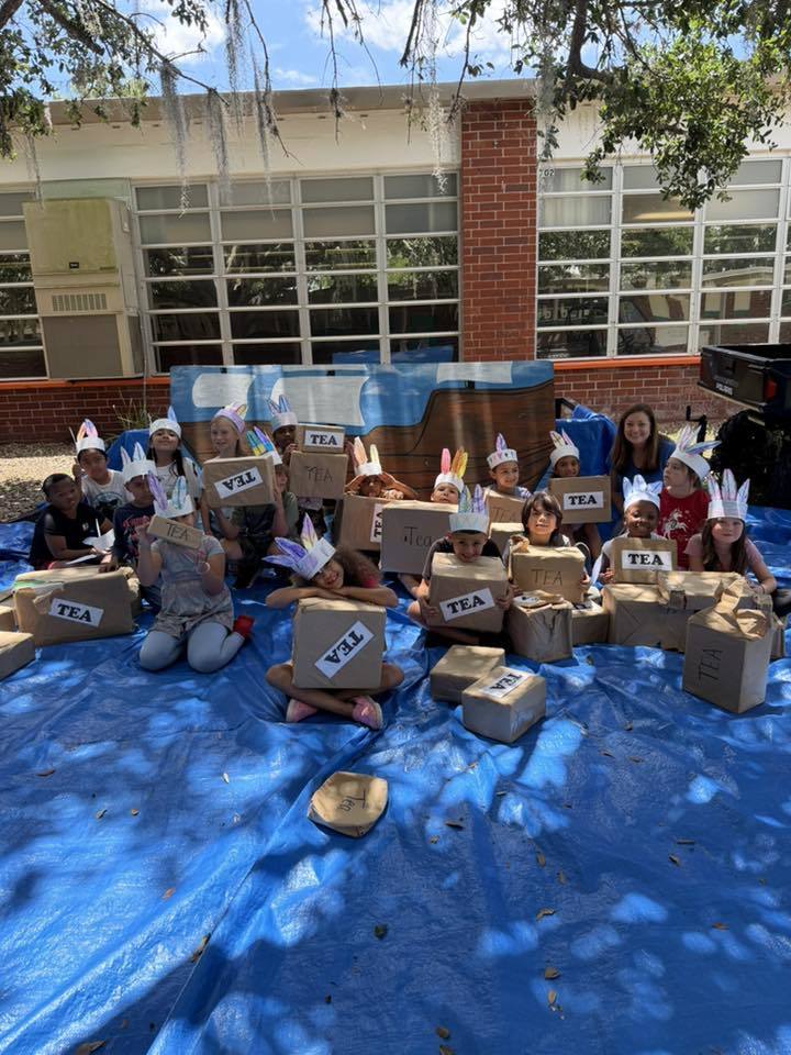 A group of elementary students sitting on a blue tarp outdoors, reenacting the Boston Tea Party. They wear feathered paper headbands and hold cardboard boxes labeled "TEA" in front of a painted backdrop of a ship.