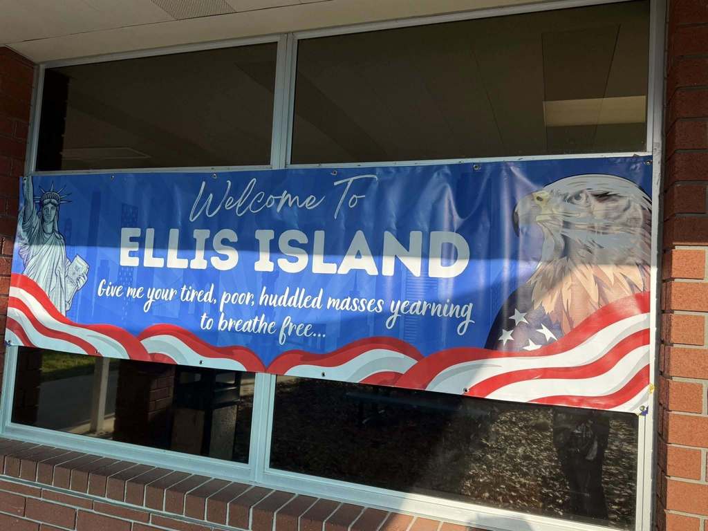 A blue vinyl banner hangs on a window, reading "Welcome To ELLIS ISLAND" with the quote "Give me your tired, poor, huddled masses..." and patriotic imagery.