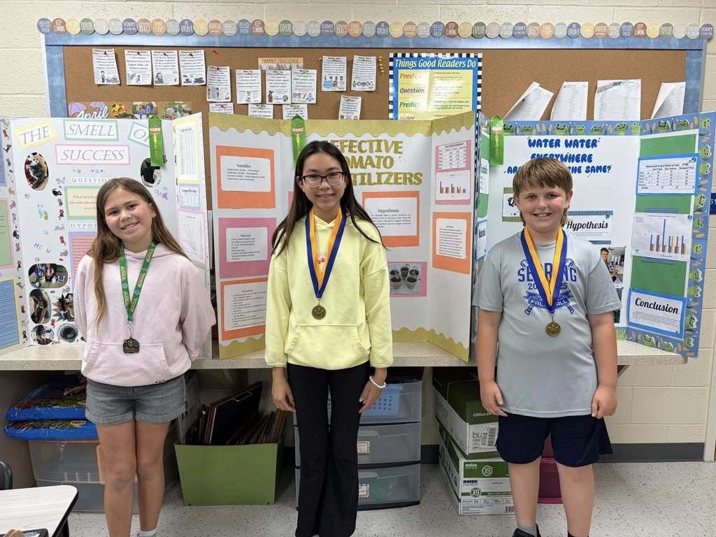 Three students stand proudly in a classroom, wearing medals around their necks in front of their science fair tri-fold displays about success, fertilizers, and water.