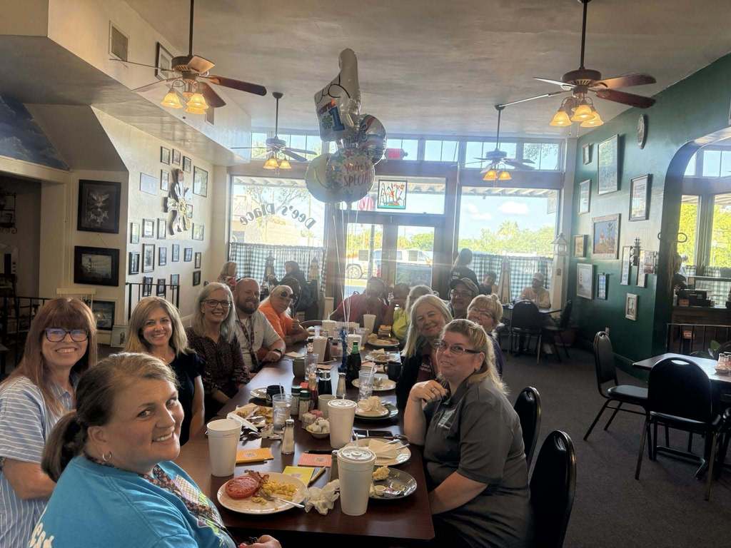 A group of adults sits around a long table in a brightly lit restaurant, smiling for a photo. Celebration balloons are visible in the background.