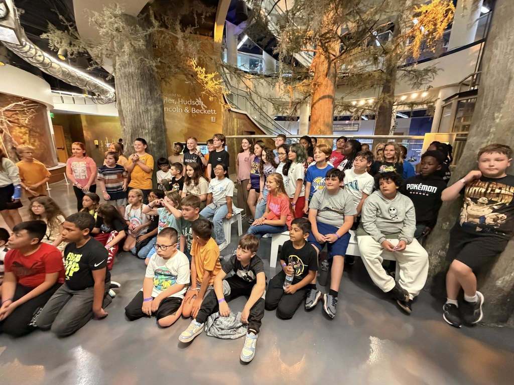 A large group of elementary-aged students sits and stands together for a photo inside a museum featuring large, artificial cypress trees and exhibit displays.