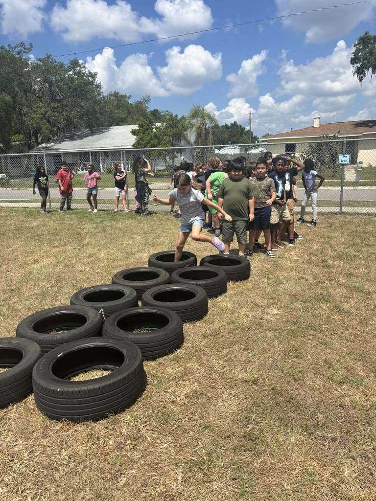 A young girl hops through a row of staggered tires on a grassy field. A line of children waits their turn behind her under a blue, cloudy sky.