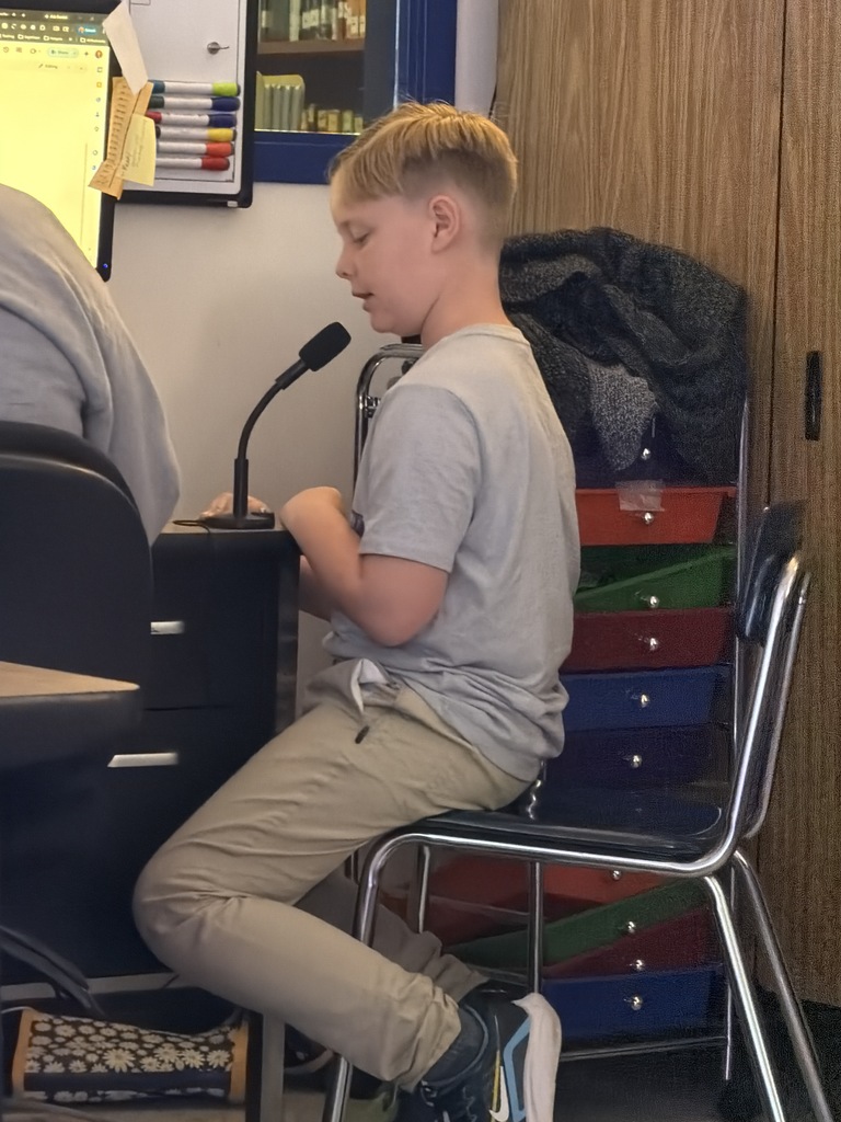 A boy with a blonde undercut hairstyle, wearing a grey t-shirt and khaki pants, sits at a desk while speaking into a microphone.
