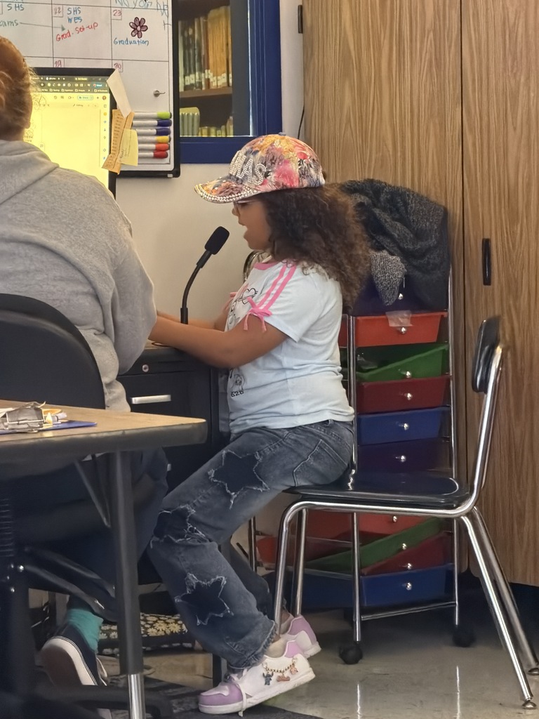 A girl with long curly hair and a floral "THOMAS" baseball cap wears a light blue t-shirt and star-patterned jeans while speaking into a microphone.
