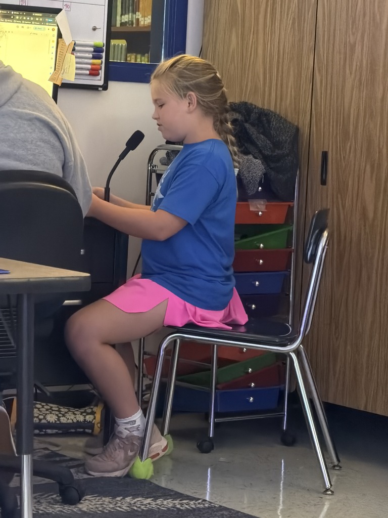 A girl with a blonde braid wearing a blue t-shirt and pink skirt sits at a desk, speaking into a microphone. The chair feet are covered with tennis balls.