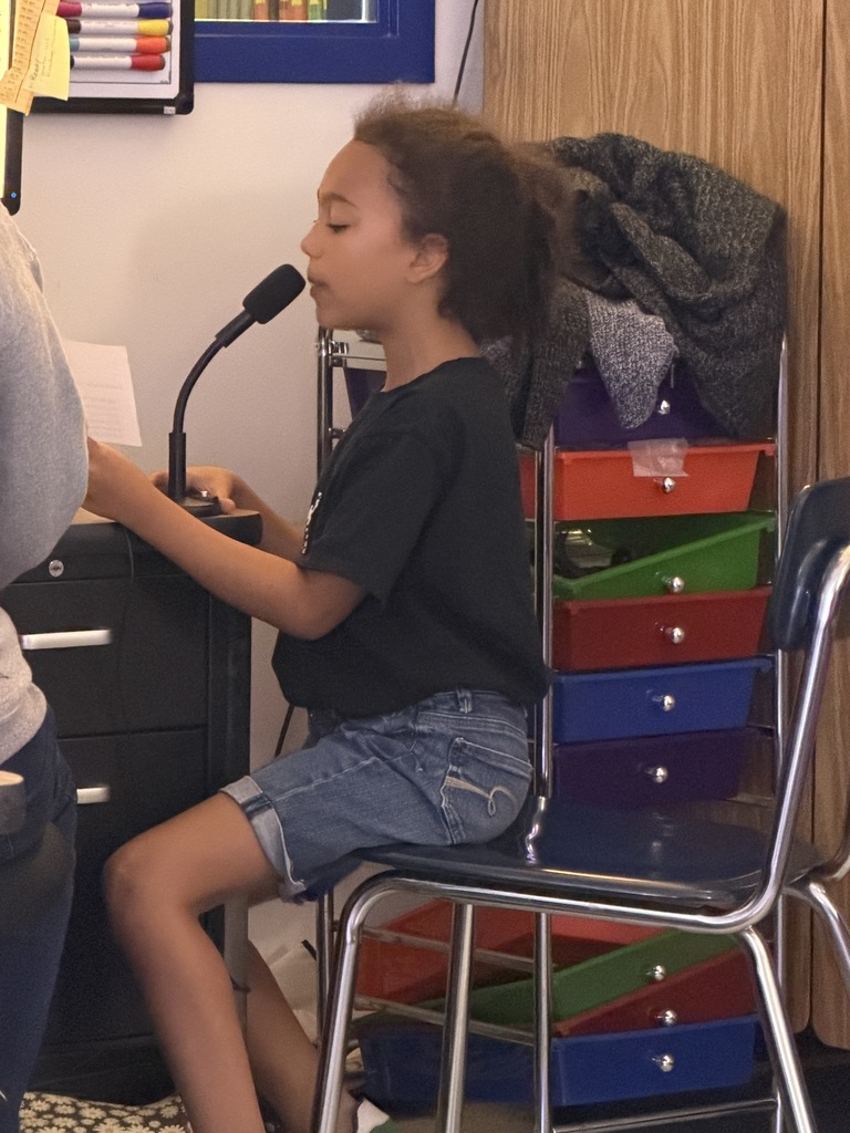 A girl in a black t-shirt and denim shorts sits on the edge of a chair, leaning toward a microphone. A stack of colorful drawers is in the background.