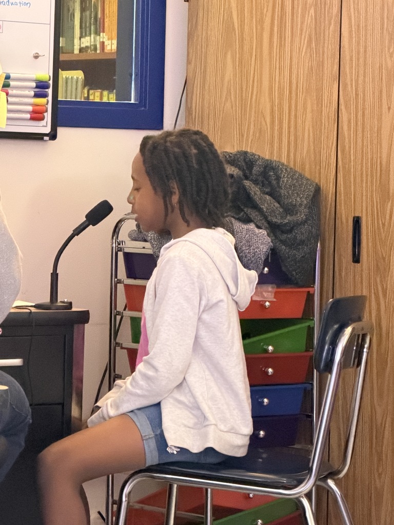 A girl with braided hair wearing a white hoodie and denim shorts sits on a blue chair, speaking into a microphone with her eyes closed in concentration.