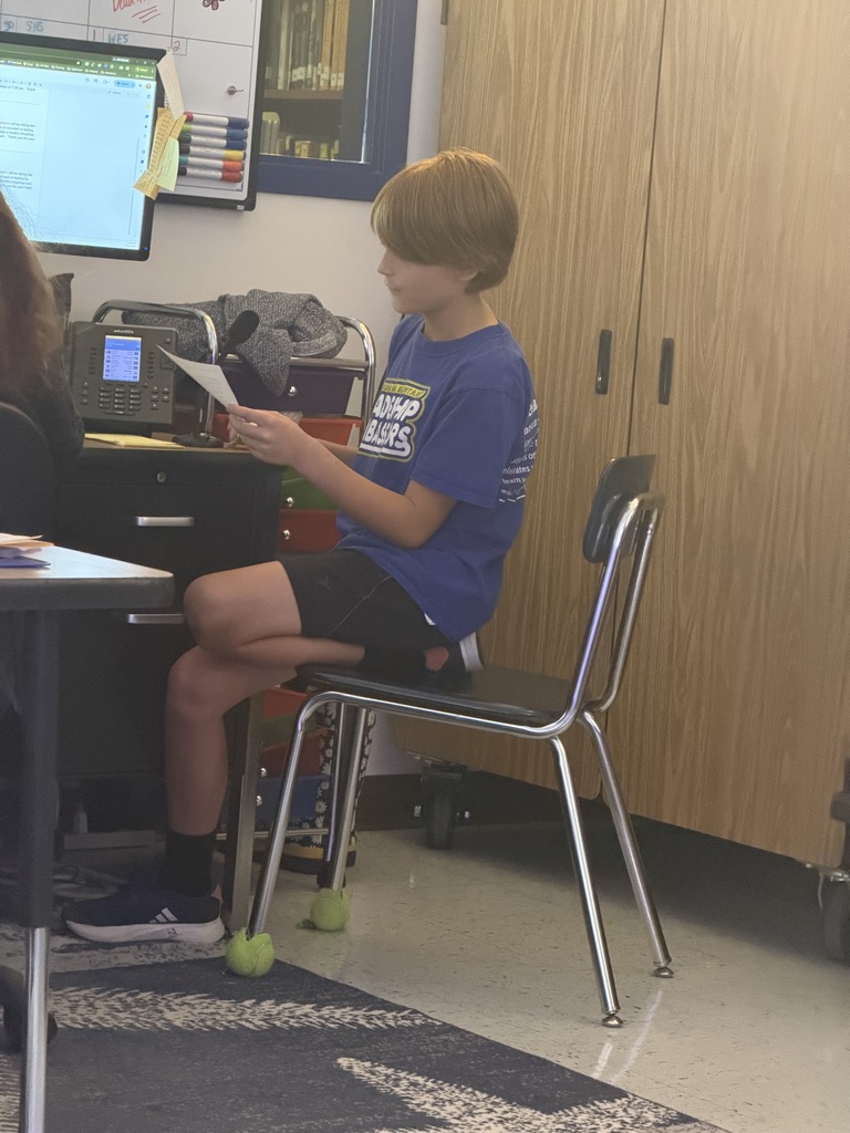 A young boy in a blue t-shirt sits on a classroom chair, holding a paper and speaking into a microphone. The chair legs have tennis balls on the feet.