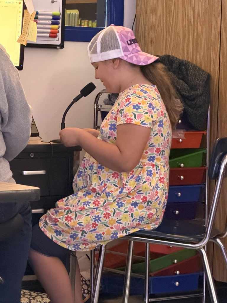 A girl in a floral dress and pink patterned baseball cap sits on a metal chair, speaking into a black gooseneck microphone. Colorful storage bins are visible behind her.
