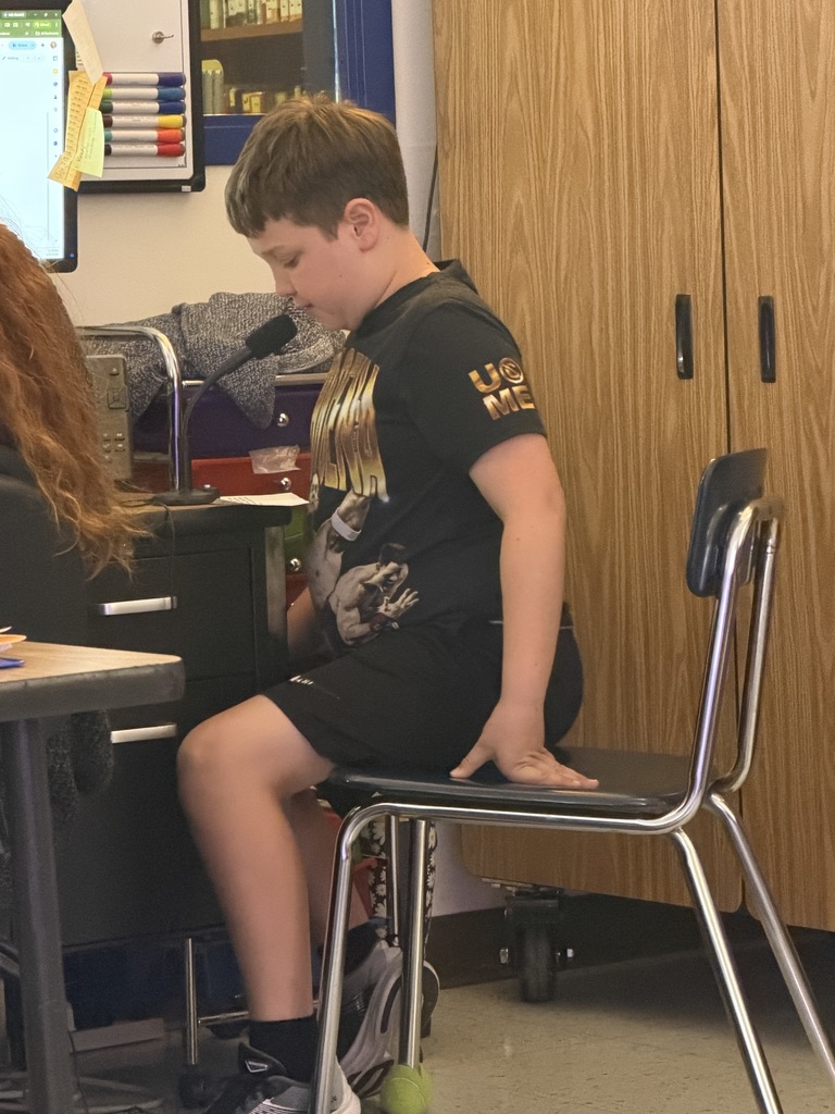 A boy wearing a black graphic t-shirt sits at a desk, leaning forward slightly to speak into a desk-mounted microphone in a classroom setting.