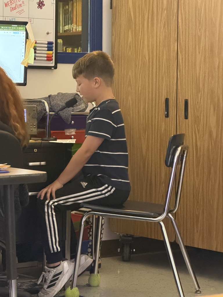 A boy in a navy striped t-shirt and Adidas track pants sits upright at a desk, speaking into a microphone. Tennis balls are attached to the front chair legs.
