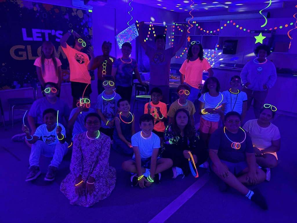 A group of approximately twenty children poses in a room illuminated by purple blacklights. They wear neon clothing and glowing accessories like necklaces, bracelets, and glasses. Festive decorations, including "LET’S GLOW" signage and hanging streamers, enhance the vibrant, neon-themed party atmosphere.