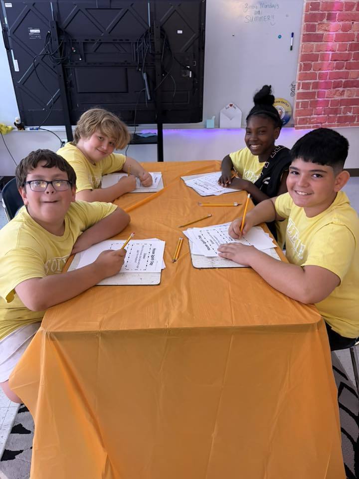 Four smiling students in matching yellow t-shirts sit around a table covered with an orange tablecloth. Each child is holding a pencil and working on a "House Spirit Day" activity sheet. In the background, a whiteboard notes there are "3 more Mondays until summer."