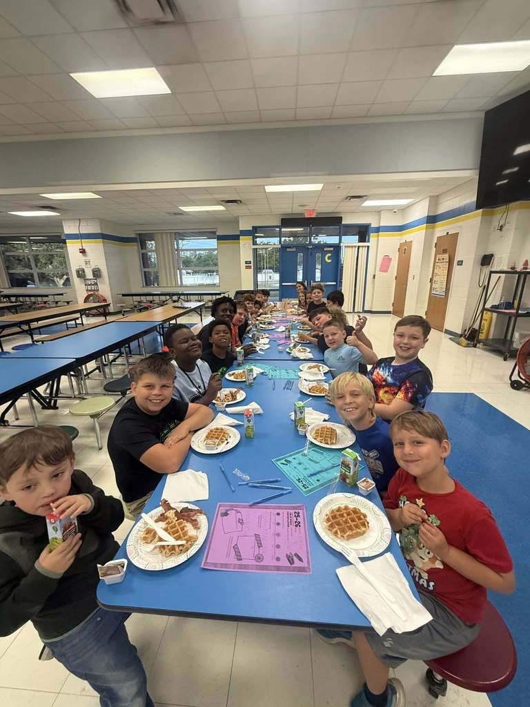 A group of approximately fifteen elementary-aged students sits at a long blue cafeteria table, smiling for the camera. Each student has a plate with a waffle and bacon, a juice box, and a purple activity sheet. The school cafeteria has white tiled walls and blue accents.