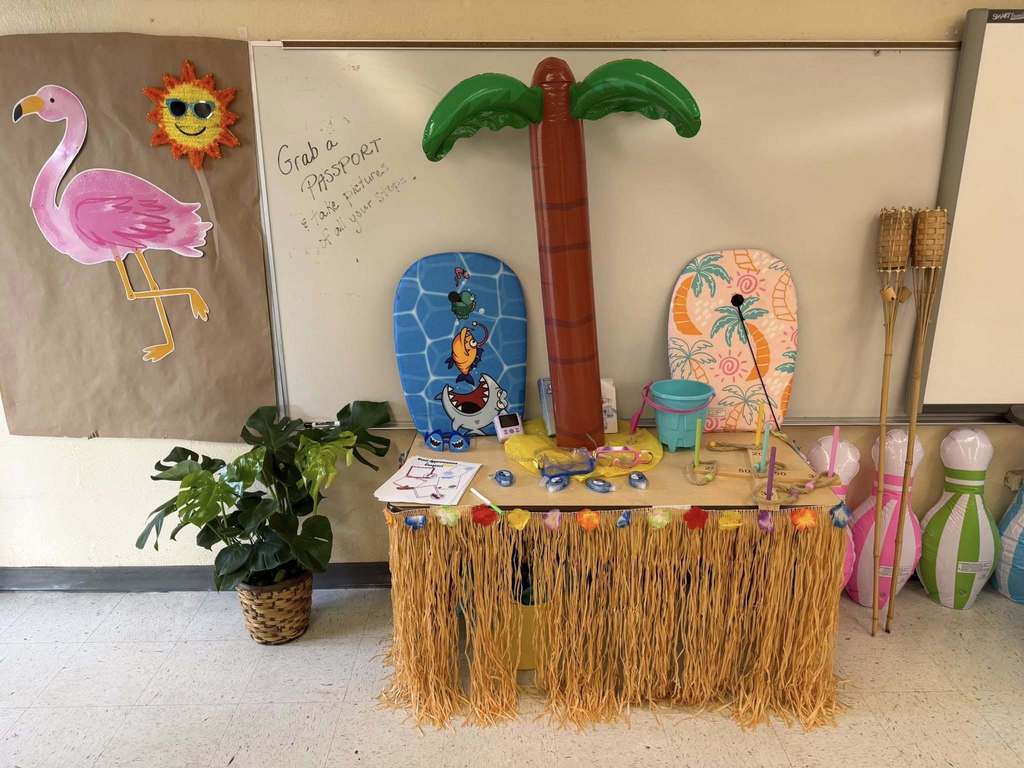 A tropical-themed classroom display features an inflatable palm tree, flamingo cutout, and grass-skirted table. Two bodyboards lean against a whiteboard labeled "Grab a Passport." Various beach accessories, including snorkels and a bucket, are arranged on the table alongside inflatable bowling pins and tiki torches.