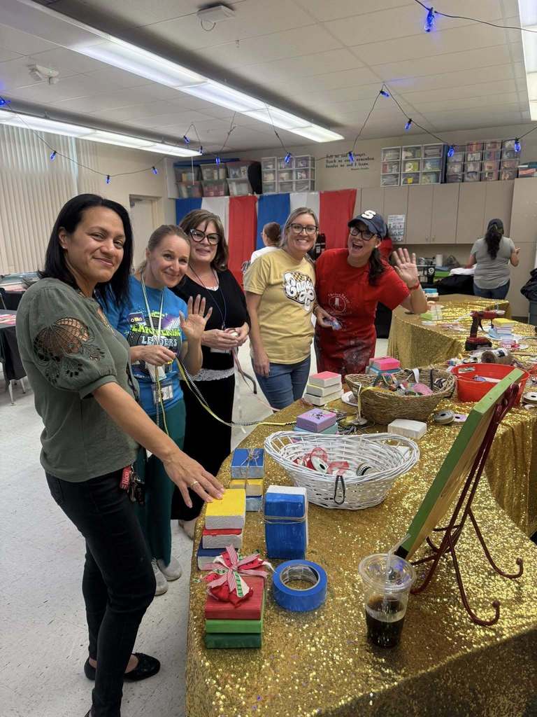  Five women smile and wave in a classroom-like setting behind a gold sequined table. They are crafting colorful wood block "books" decorated with twine and ribbon. Blue string lights hang overhead, and red, white, and blue banners line the back wall. Baskets and craft supplies are scattered across the table.