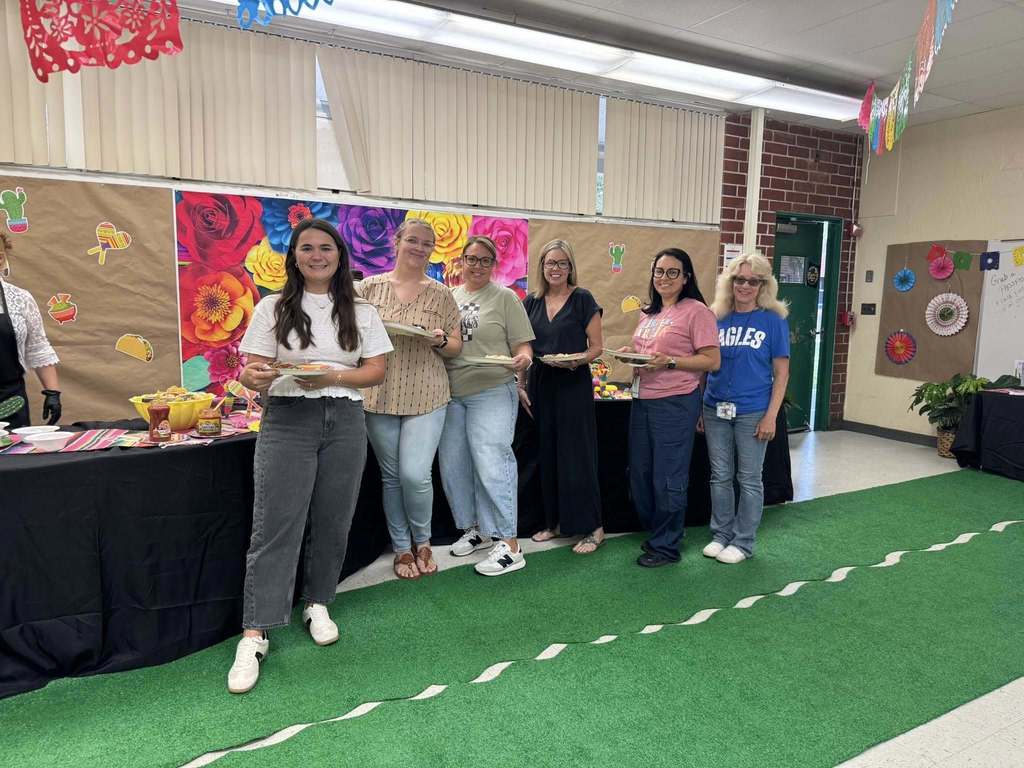 Six women stand in a decorated classroom during a festive event. They are lined up behind a table with food, several holding plates. The room features a colorful floral backdrop, taco-themed decorations, and green turf on the floor. Most of the women are smiling and wearing casual attire.