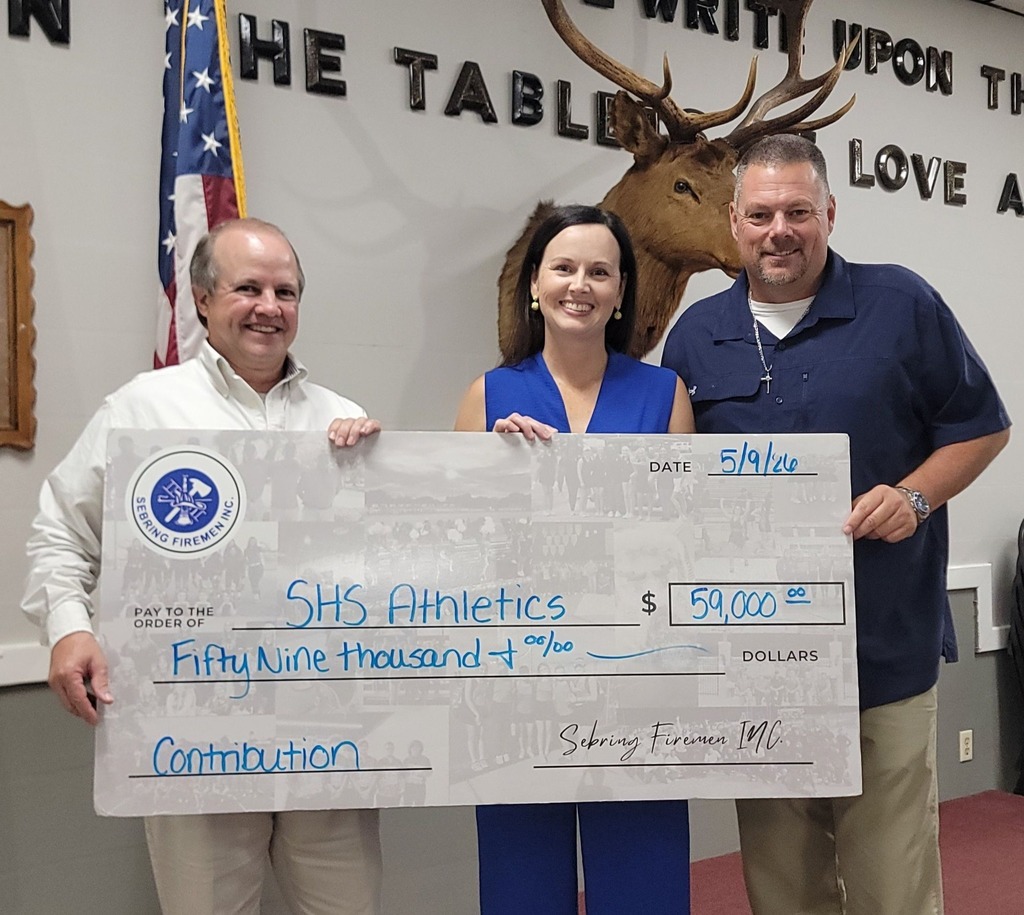 Three people stand smiling behind a large ceremonial check for $59,000. The check is made out to SHS Athletics from Sebring Firemen Inc., dated 5/9/26. An American flag and a mounted elk head are visible on the wall behind them.