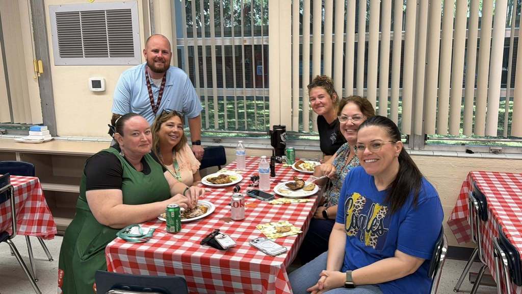 A group of six smiling adults gathers around a table with a red-and-white checkered tablecloth in a brightly lit room. They are enjoying a meal of grilled chicken and sides. The setting includes large windows with blinds and classroom-style furniture, suggesting a school or community event.