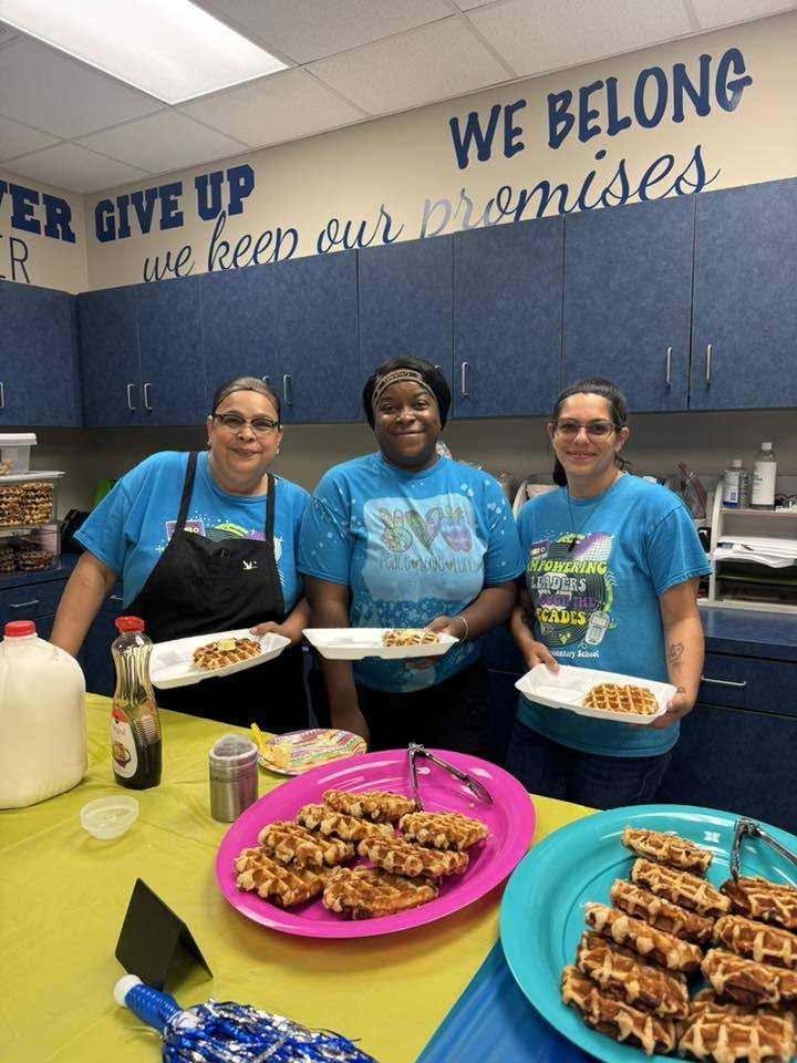  Three women in blue shirts stand behind a table filled with waffles, each holding a served plate. The background features blue cabinetry and wall text reading, "WE BELONG" and "we keep our promises." A large pink platter and a blue platter in the foreground display several golden-brown waffles.