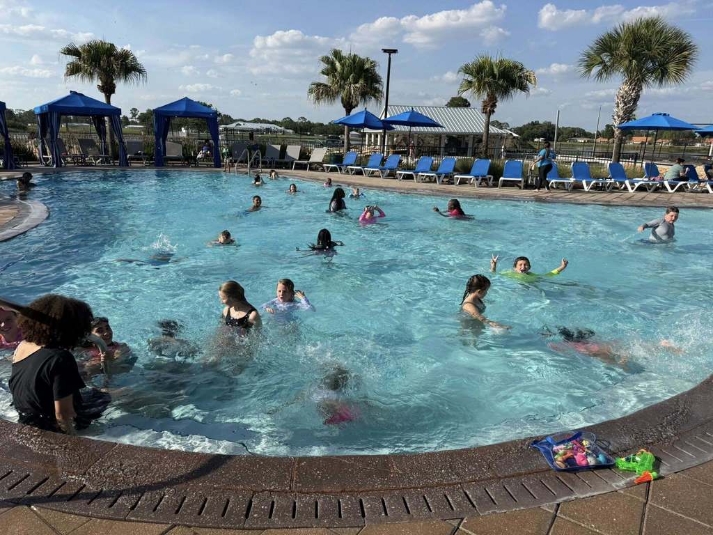 A vibrant outdoor swimming pool filled with children playing under a clear, sunny sky. Palm trees, blue cabanas, and lounge chairs line the pool deck. The pool features a curved edge with several kids swimming, splashing, and wading in the turquoise water, creating an active summer scene.