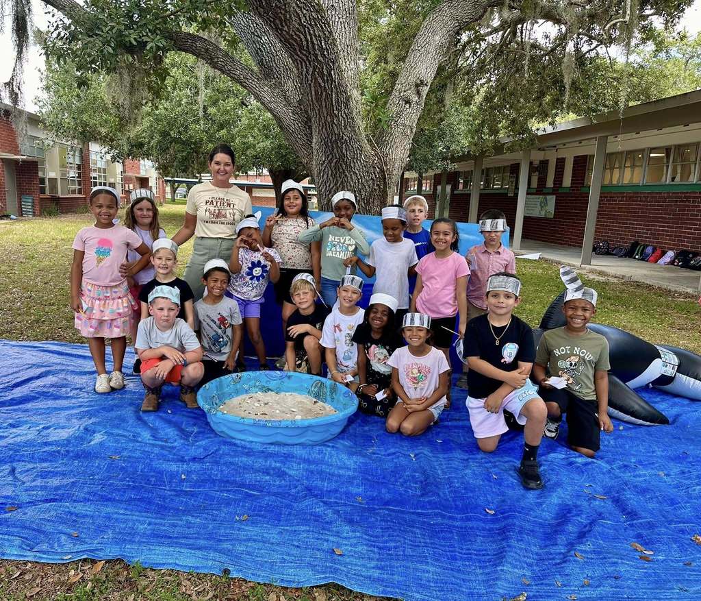 A group of approximately twenty young students and their teacher pose outdoors on a large blue tarp. The children wear handmade paper headbands, some featuring shark fins. They are gathered around a small blue pool filled with sand, positioned in front of a large tree and an inflatable shark.
