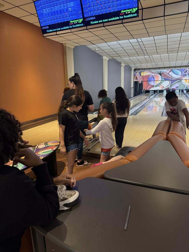 A group of children and adults are gathered at a bowling alley. Several people stand near the ball return, while a young boy prepares to bowl in the background. An overhead electronic scoreboard displays player scores. The foreground shows a seating area with a table and a pen.