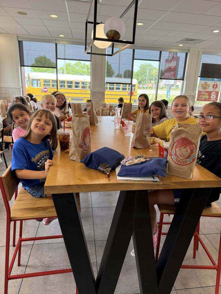  A group of children sit smiling around a long wooden table inside a Wendy's restaurant. They have Wendy's brown paper bags and drinks in front of them. The table features large, black, W-shaped legs. Outside the large windows, a yellow school bus is visible.