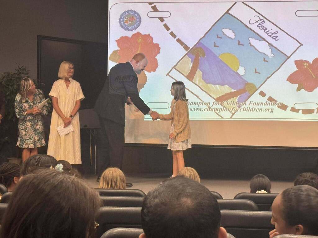 A young girl in a cardigan shakes hands with a man in a suit on a stage. Behind them, a large screen displays her colorful artwork of a Florida beach scene framed as a license plate. Two women watch from the side.
