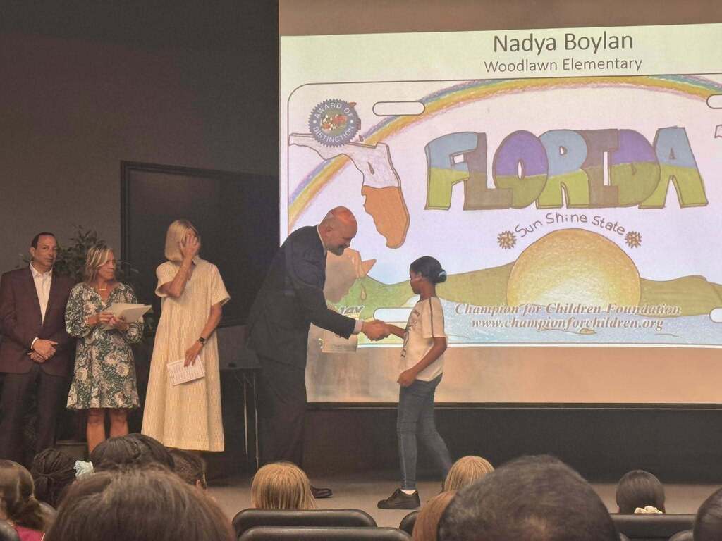 On a stage, a man in a suit shakes hands with a student named Nadya Boylan from Woodlawn Elementary. The large screen behind them displays her colorful rainbow and sunshine license plate artwork. Three adults stand on the left.