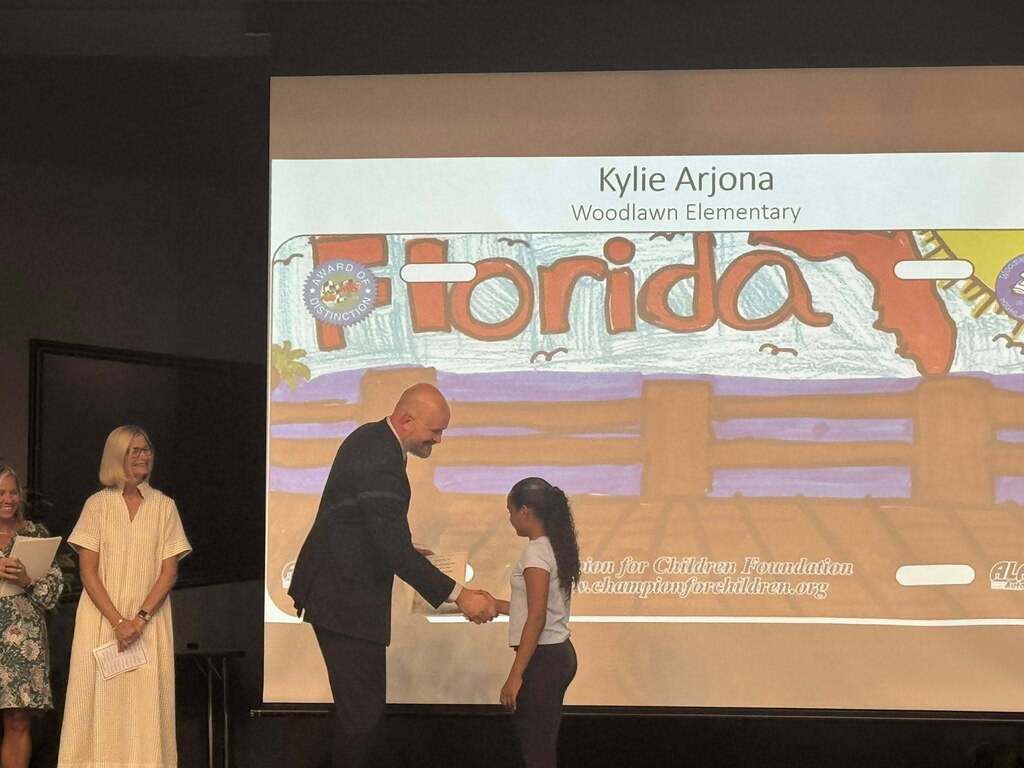 A student named Kylie Arjona from Woodlawn Elementary shakes hands with a man on stage. Behind them, a large screen shows her artwork featuring a wooden pier and the word "Florida" stylized as a license plate. Two women smile from the left side.