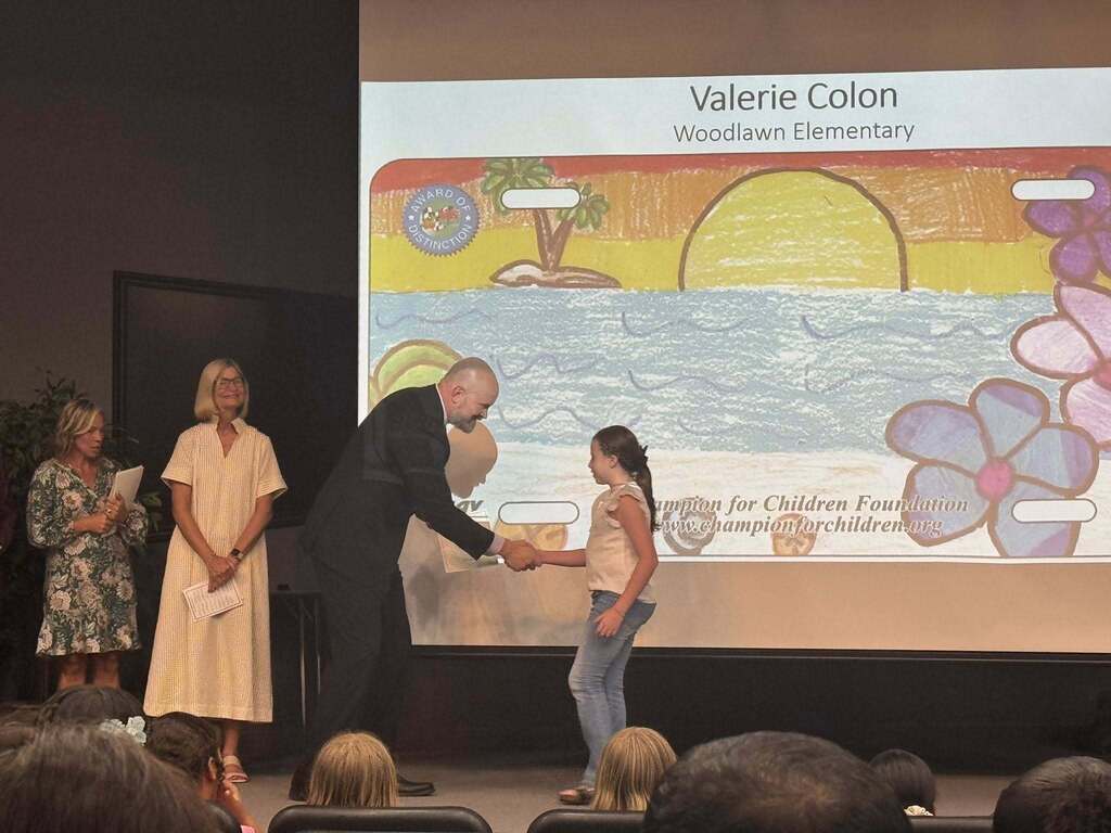 A young girl named Valerie Colon from Woodlawn Elementary shakes hands with a man in a suit on a stage. The background screen displays her vibrant drawing of a tropical beach sunset designed like a Florida license plate. Two women watch nearby.