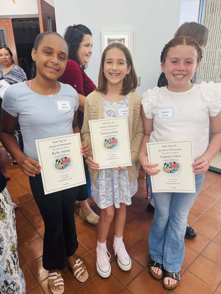 Three smiling young girls stand side-by-side indoors, wearing name tags and holding "Kids Tag Art Award of Distinction" certificates. From left to right, their name tags read Kylie Arjona, Emerson Bury, and Valerie Colon. Adults stand in the background.