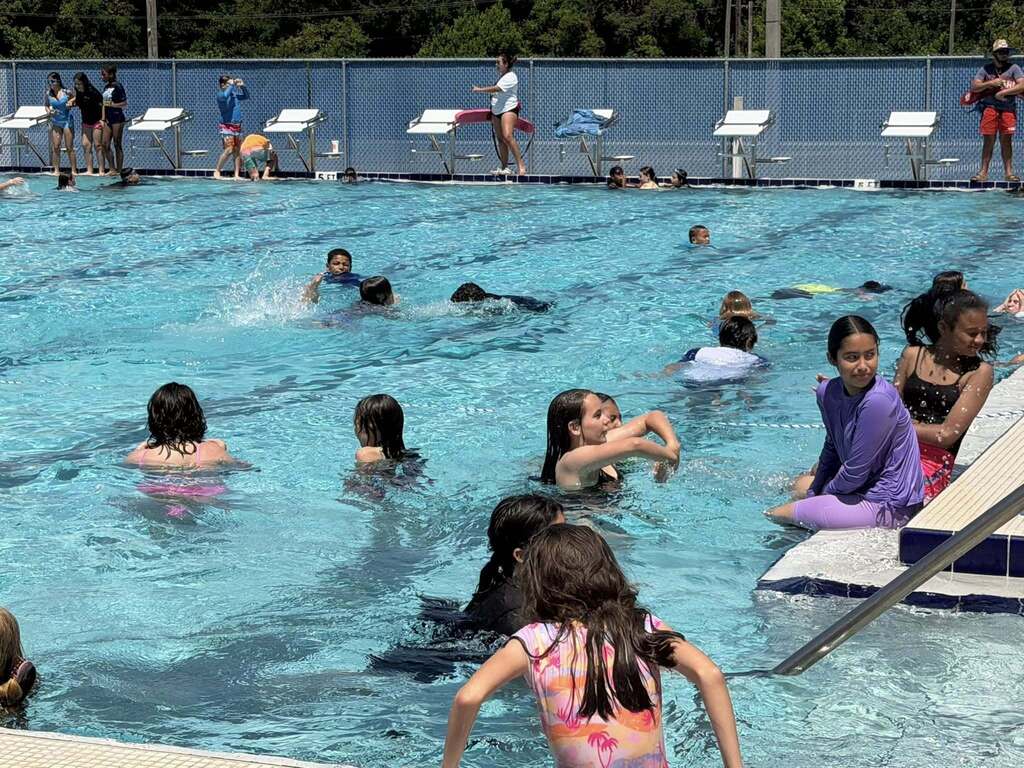  A crowded outdoor swimming pool filled with children and teens swimming and playing. Several people sit along the concrete pool edge, while others stand near diving blocks along the far side fence. Lifeguards are visible on the pool deck. The water is bright blue under clear daylight.