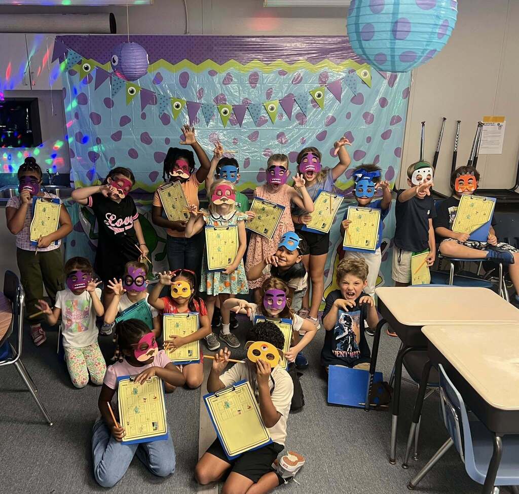  A group of around fifteen smiling young children pose in a classroom, wearing colorful, handmade monster masks. Most hold up yellow clipboards, and several make playful "monster" hand gestures. They are gathered in front of a blue and purple spotted backdrop adorned with paper lanterns and monster-eye bunting.