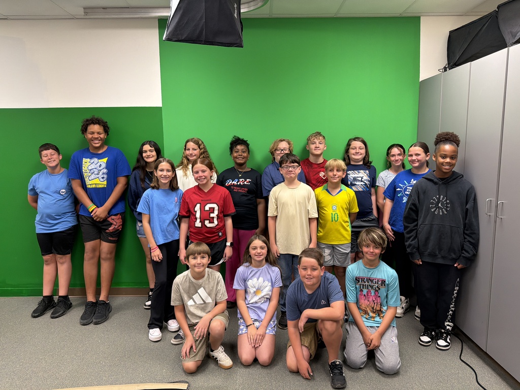 A large group of diverse school-aged children pose together for a group photo in front of a bright green screen background. Four children kneel in the front row, while the rest stand in a line behind them, smiling at the camera.